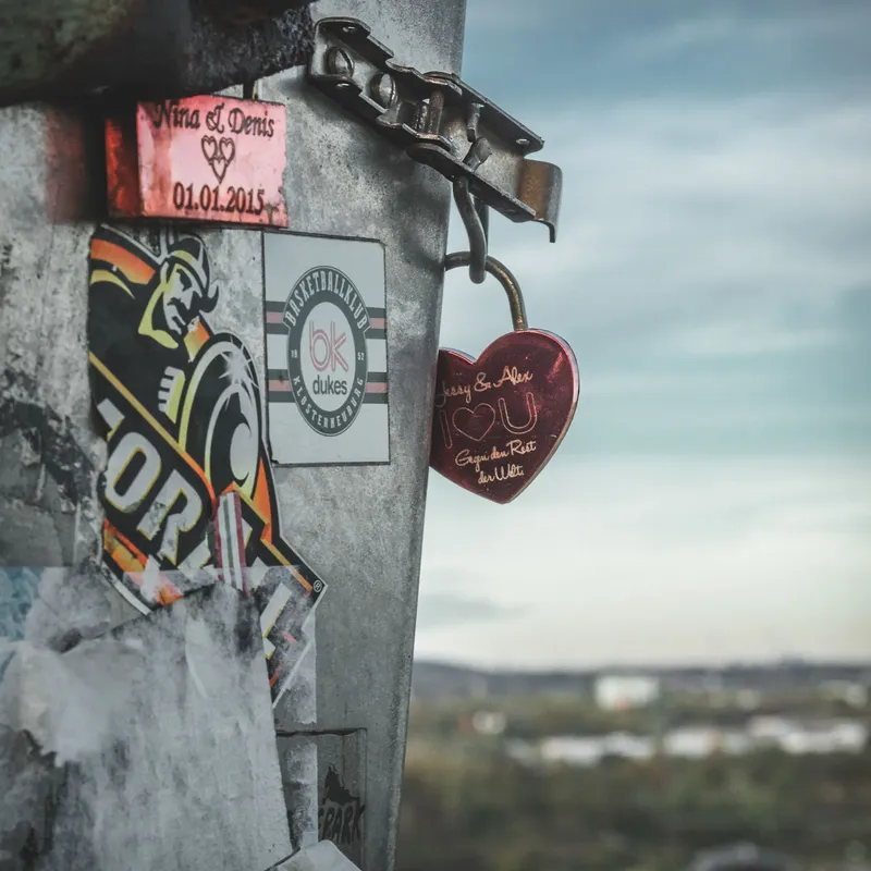 Atmosphärisches Foto eines roten Liebesschlosses an einer Aussichtsplattform mit Stadtpanorama. Romantische Szene mit gravierten Herzen, urbanen Stickern und dem Datum 01.01.2015 vor einem stimmungsvollen Himmel. Liebesschloss Stadtansicht Romantik
