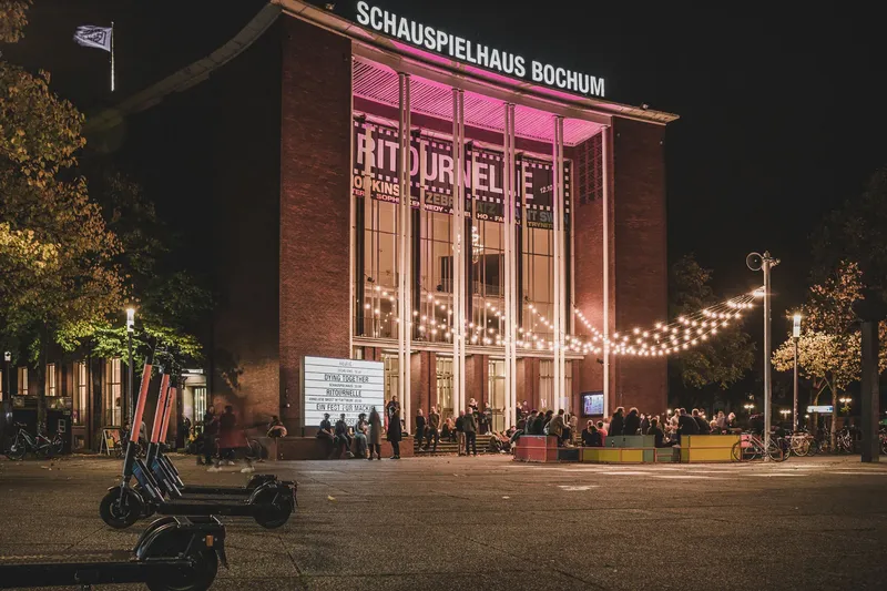 Atmosphärische Nachtaufnahme des Schauspielhaus Bochum mit pinker Beleuchtung, historischer Backsteinfassade und lebendigem Theaterpublikum im Eingangsbereich. Das kulturelle Herzstück der Ruhrmetropole präsentiert sich mit stimmungsvoller Außenbeleuchtung und modernem urbanen Flair.
