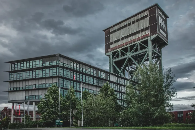 Imposante Industriearchitektur in Deutschland: Modernes Bürogebäude mit charakteristischem Förderturm vor dramatischem Wolkenhimmel. Grüne Parkanlage und zeitgenössische Architektur vereinen industrielles Erbe mit modernem Design. Perfekte Symbiose aus Vergangenheit und Gegenwart.
