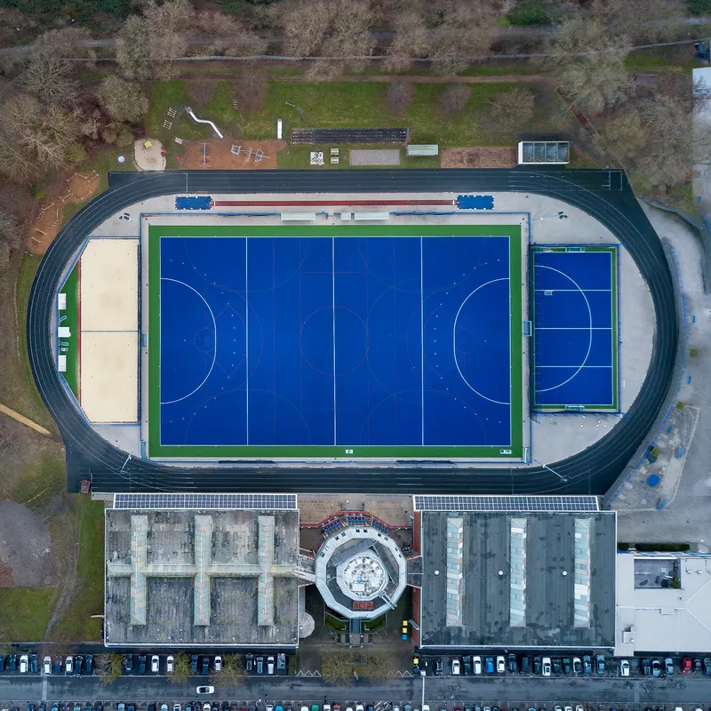 Moderne Sportanlage aus der Vogelperspektive: Professioneller blauer Kunstrasenplatz mit Wettkampf-Laufbahn, Trainingsfeld und Sportgebäude. Ideale Sport- und Trainingseinrichtung für Schul- und Vereinssport. Hochwertige Drohnenaufnahme zeigt die komplette Infrastruktur der Anlage.

