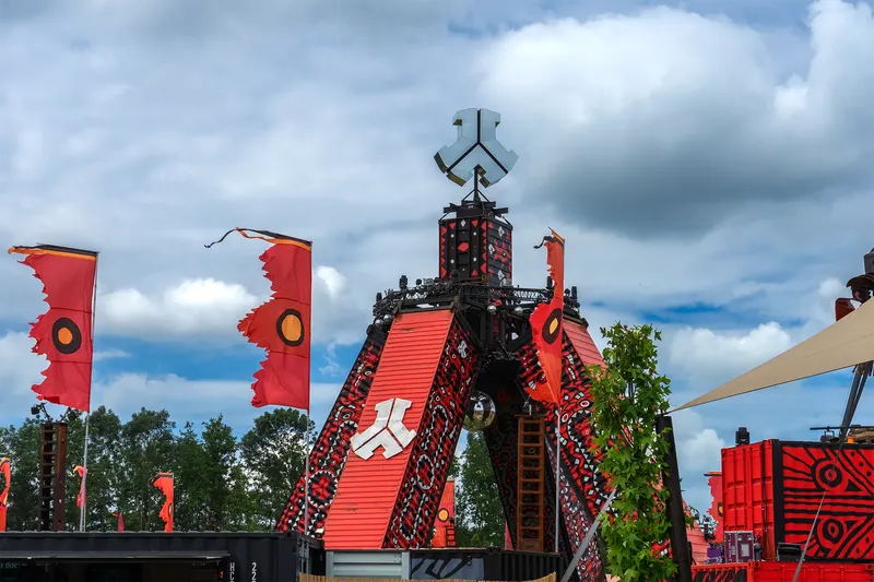 Imposante Festival-Installation mit roter A-förmiger Hauptstruktur, tribalen Mustern und wehenden Bannern vor blauem Himmel. Industrial-Style Architektur kombiniert mit moderner Festivalästhetik. Professionelle Event-Fotografie in hoher Auflösung (1536x1024).
