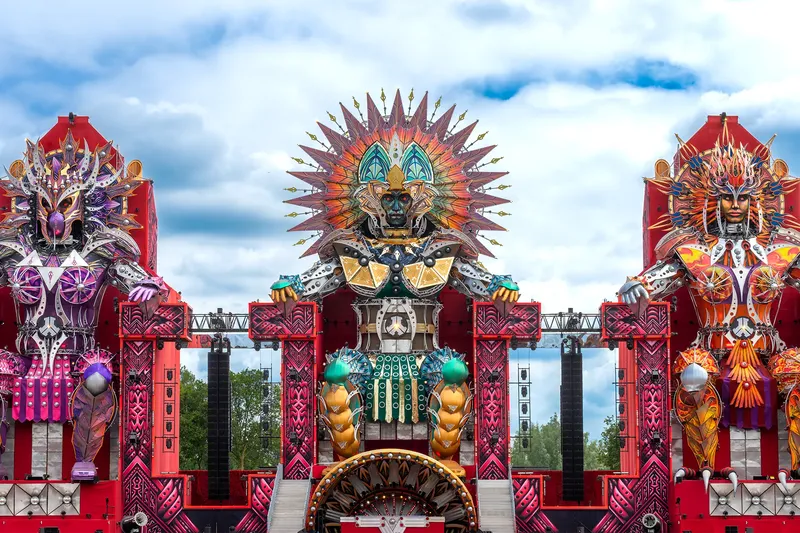 Spektakuläre EDM Festival Bühneninstallation mit drei imposanten Tribal-Skulpturen in futuristischem Design. Kunstvolle Verschmelzung von aztekischer Mythologie und moderner Festivalkultur in beeindruckender Größe mit aufwändiger Beleuchtung und geometrischen Mustern in kräftigen Farben.
