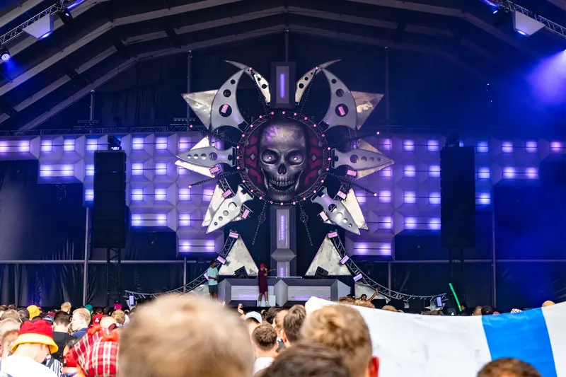 Eindrucksvolle Festival-Bühne mit imposanter Totenkopf-Installation, umgeben von futuristischen Metallelementen und atmosphärischer blau-violetter Lichtshow. Moderne EDM-Festival-Atmosphäre mit begeistertem Publikum vor beeindruckender Bühnentechnik und innovativem Stage-Design.
