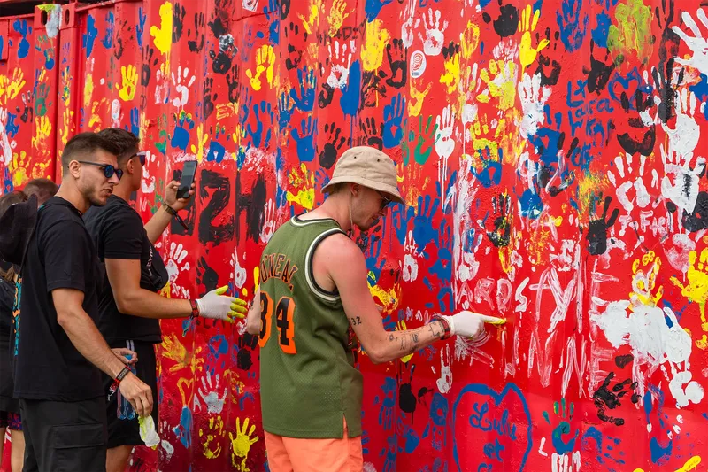 Lebendige Gemeinschaftskunst: Bunte Handabdrücke auf roter Wand zeigen urbane Kreativität und partizipative Straßenkunst. Ein dynamisches Kunstprojekt mit Handprints in Gelb, Blau und Weiß demonstriert gemeinschaftliches Engagement in der Street-Art-Szene.
