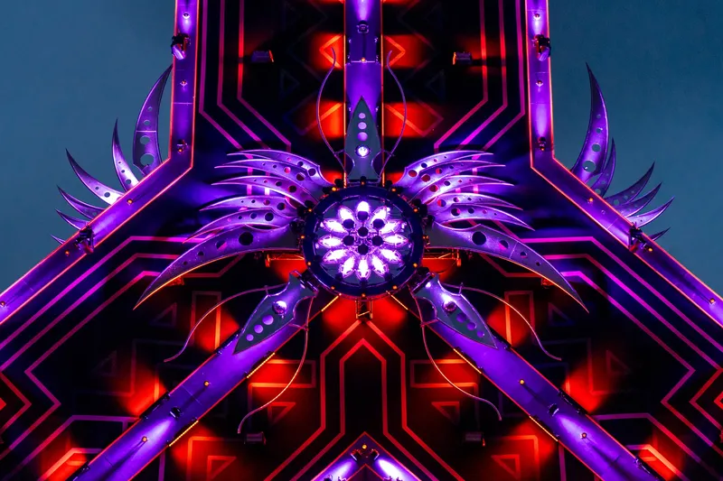 Futuristische Lichtinstallation in leuchtendem Violett und Rot mit geometrischem Design. Zentrale Mandala-ähnliche Struktur mit LED-Beleuchtung, umgeben von symmetrischen Metallstrukturen. Perfekt für elektronische Musikfestivals, Bühnenshows und moderne Kunstinstallationen.
