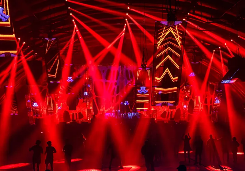 Spektakuläre Nachtclub-Atmosphäre mit imposanter Lichtshow in intensivem Rot, futuristischer LED-Installation und geometrischen Bühnenelementen. Professionelle Event-Fotografie zeigt dynamische Festival-Stimmung mit Silhouetten tanzender Menschen unter beeindruckenden Moving-Lights und modernem Bühnendesign.
