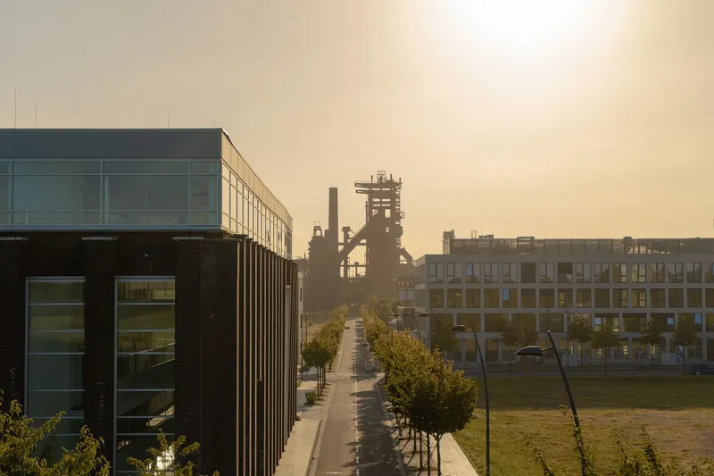 Moderne Industriearchitektur trifft auf urbanes Design: Imposante Industrieanlage mit Hochofen im goldenen Abendlicht, flankiert von zeitgenössischen Bürogebäuden und einer baumgesäumten Allee - ein eindrucksvolles Beispiel für gelungene Industriekultur und nachhaltige Stadtentwicklung.
