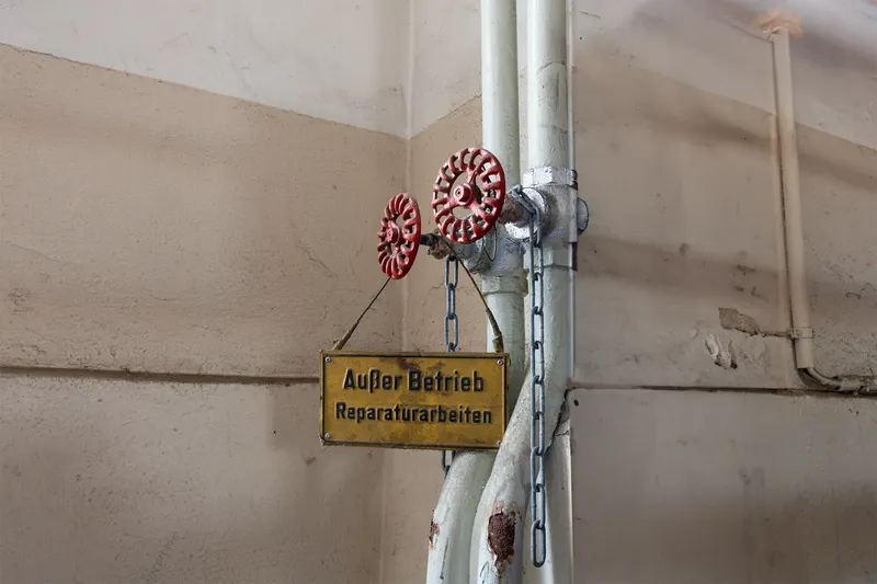 Industrielle Wartungsankündigung mit gelbem 'Außer Betrieb' Warnschild und roten Ventilrädern an grauer Rohrleitung. Dokumentation von Reparaturarbeiten und Industriesicherheit im deutschsprachigen Raum.
