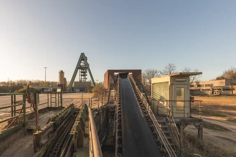 Industrielle Förderanlage einer Bergbaueinrichtung mit markanter A-förmiger Förderturm-Konstruktion, Förderbändern und technischen Installationen bei Sonnenaufgang. Authentische Industriearchitektur dokumentiert das kulturelle Erbe des deutschen Bergbaus.

