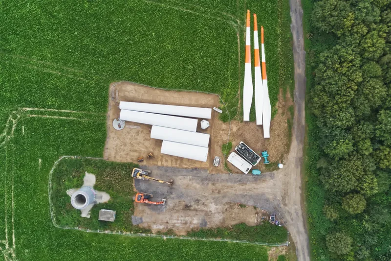 Luftaufnahme einer Windkraftanlagen-Baustelle mit gelagerten Rotorblättern und Turmsegmenten. Dokumentation der nachhaltigen Energiewende mit professionellen Drohnenaufnahmen aus der Vogelperspektive. Einblicke in den Aufbau moderner Windenergieanlagen.
