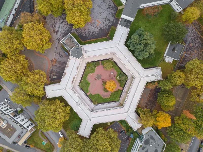 Eindrucksvolle Luftaufnahme des Leibnizgymnasiums im Herbst: Charakteristischer hexagonaler Grundriss mit weißem Dach, umgeben von herbstlich gefärbten Bäumen und gepflegten Grünanlagen. Das architektonische Wahrzeichen zeigt einen symmetrischen Innenhof mit rotem Pflaster.
