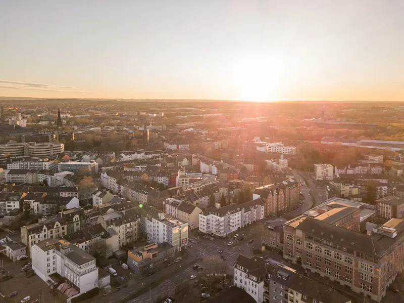 Atmosphärische Luftaufnahme der Ruhrgebietsstadt Bochum im goldenen Abendlicht. Urbane Stadtlandschaft mit historischen Gründerzeithäusern, modernen Wohngebäuden und charakteristischer Ruhrgebietsarchitektur im Panoramaformat. Authentischer Einblick in die lebendige Metropole des Ruhrgebiets.
