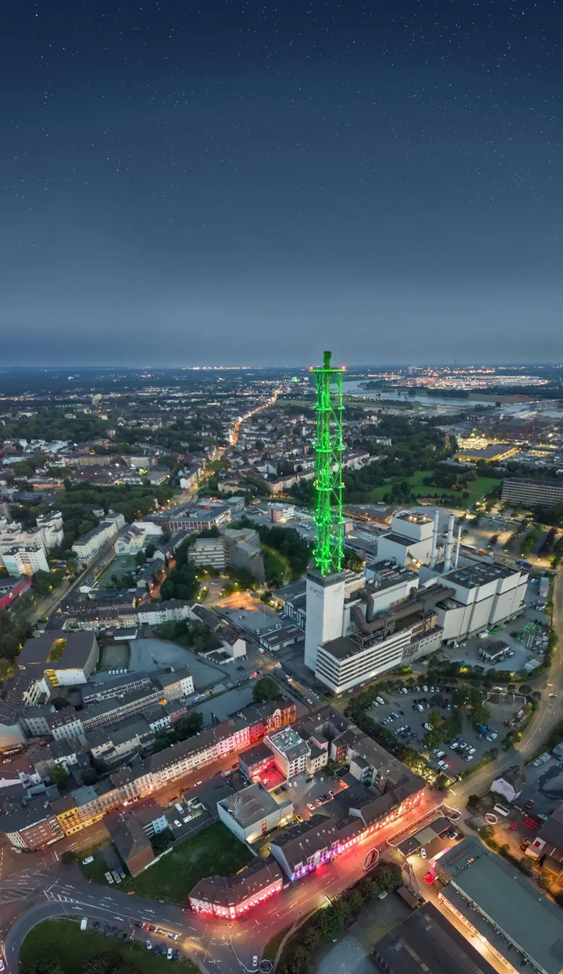Spektakuläre Nachtaufnahme des grün illuminierten Stadtwerketurms in Duisburg aus der Vogelperspektive. Das Wahrzeichen erstrahlt vor einem Sternenbesetzten Nachthimmel über der beleuchteten Ruhrgebietsmetropole und zeigt die eindrucksvolle urbane Architektur der Stadt.
