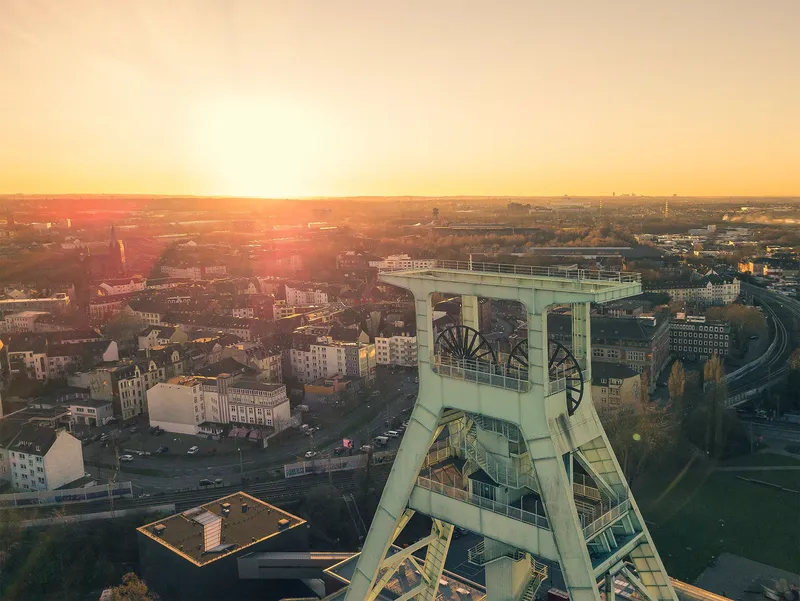 Imposantes Fördergerüst des Deutschen Bergbaumuseums Bochum im goldenen Abendlicht. Luftaufnahme zeigt historisches Industriedenkmal über der Ruhrgebietsmetropole mit charakteristischer Stadtsilhouette. Authentisches Wahrzeichen der Bergbautradition.
