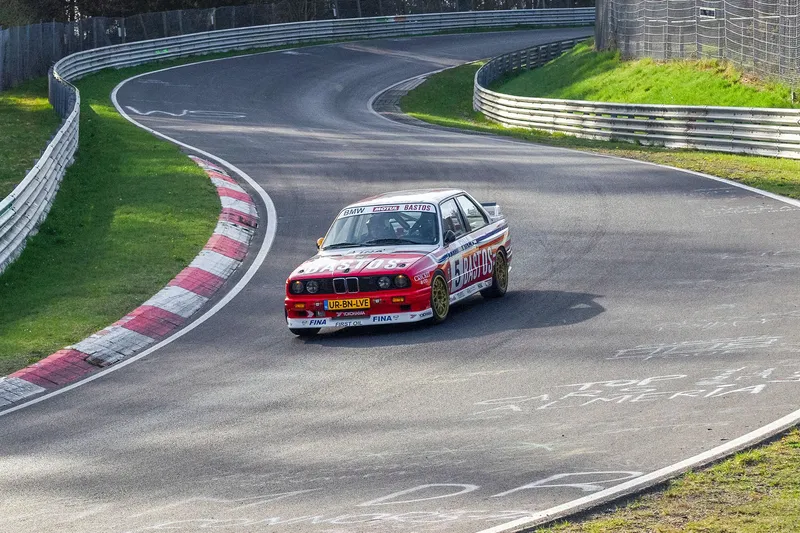 Historischer BMW-Rennwagen mit Bastos-Lackierung auf der legendären Nürburgring-Rennstrecke in Aktion. Dynamische Aufnahme zeigt die charakteristischen Kurven und Streckenbegrenzungen der weltberühmten Rennstrecke.
